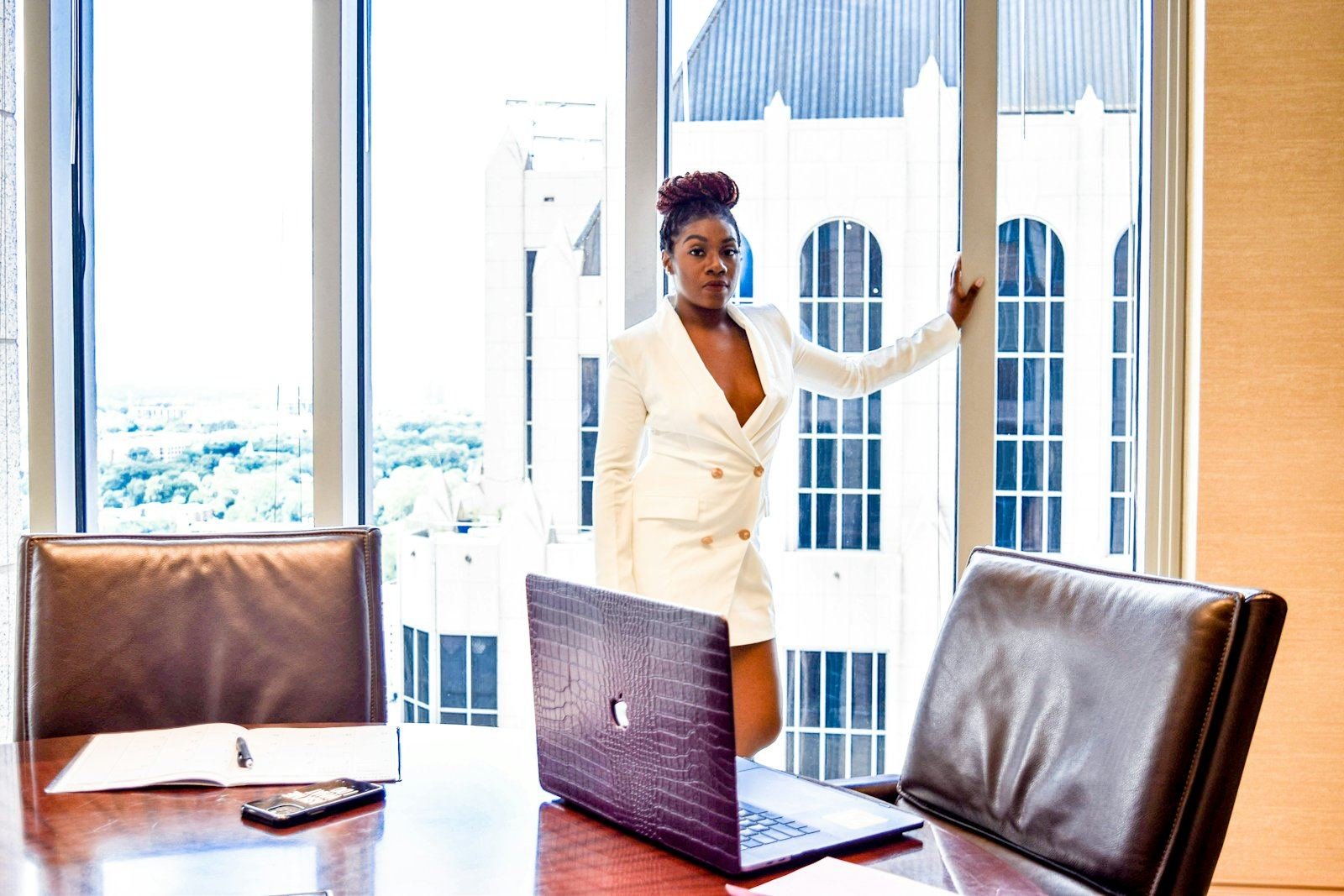 An executive in a white suit beside tall office windows, a laptop on the boardroom table