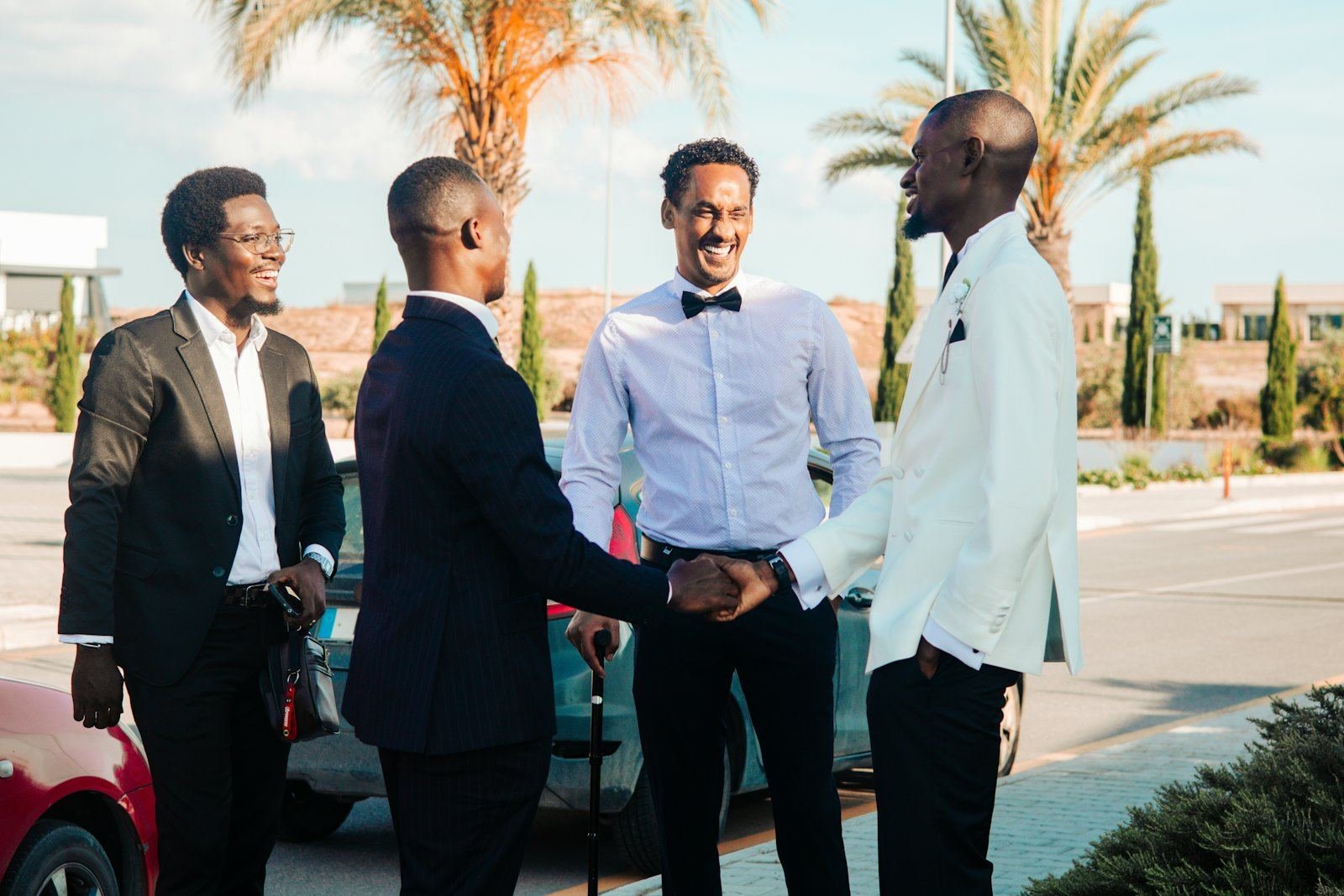 Four businessmen in suits greeting one another outdoors, smiling as two shake hands